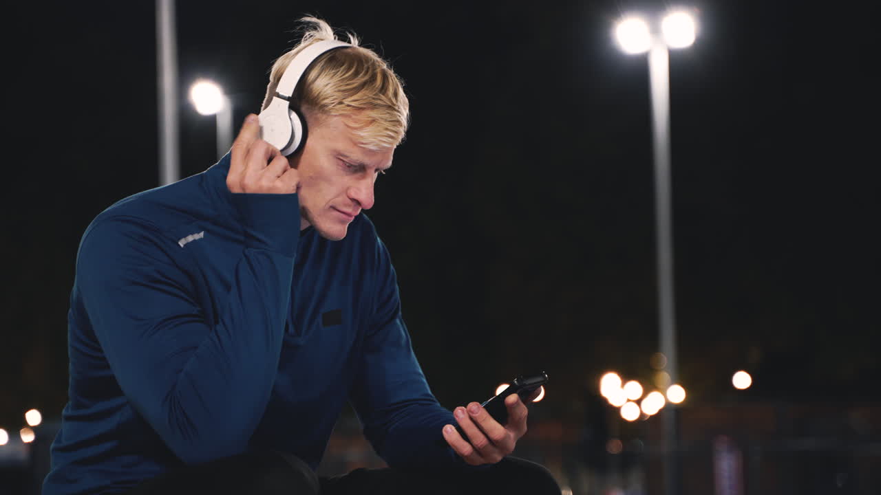 deportista sonriente sentado en el parque escuchando música con auriculares bluetooth y enviando mensajes de texto en su teléfono móvil mientras toma un descanso durante su sesión de entrenamiento por la noche
