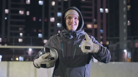 smiling athlete wearing hoodie and gloves holding soccer ball while giving thumbs up during nighttime practice session at an urban sports field, illuminated city buildings and bokeh lights