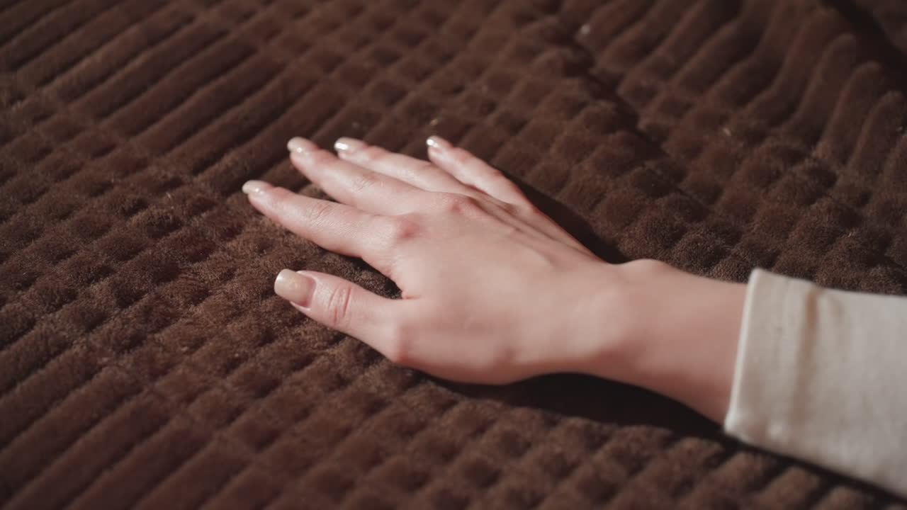 close up of female hand with polished nails touching textured brown fabric on bed, capturing gentle motion and surface feel, showing calm sensory moment inside warmly lit room interior