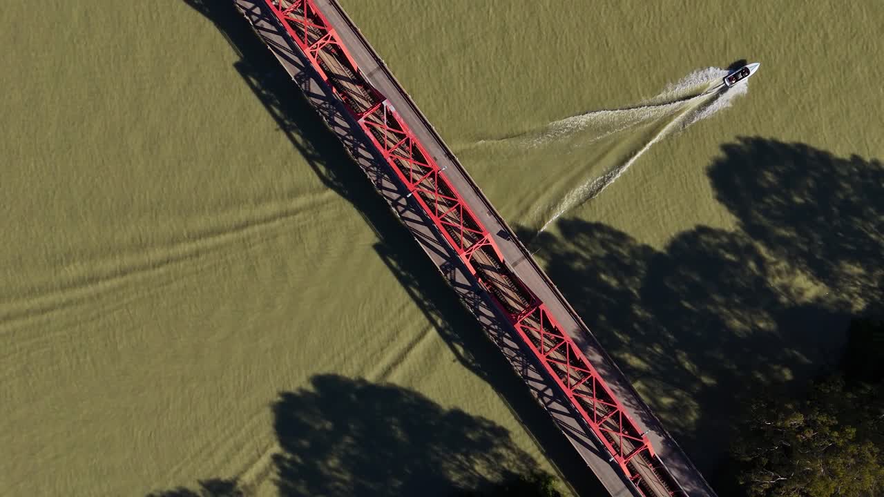 Struttura del ponte Paringa che si estende sulla superficie di un fiume verde con una forte forma lineare e ombre, vista aerea angolata dall'alto verso il basso con una barca che lo attraversa