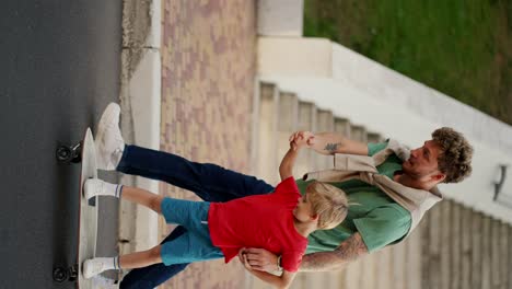 Vertical-video-of-a-dad-with-curly-hair-in-a-Green-T-shirt-and-blue-jeans-helping-his-little-blond-son-in-a-red-T-shirt-and-shorts-ride-a-white-skateboard-along-the-road-in-the-park