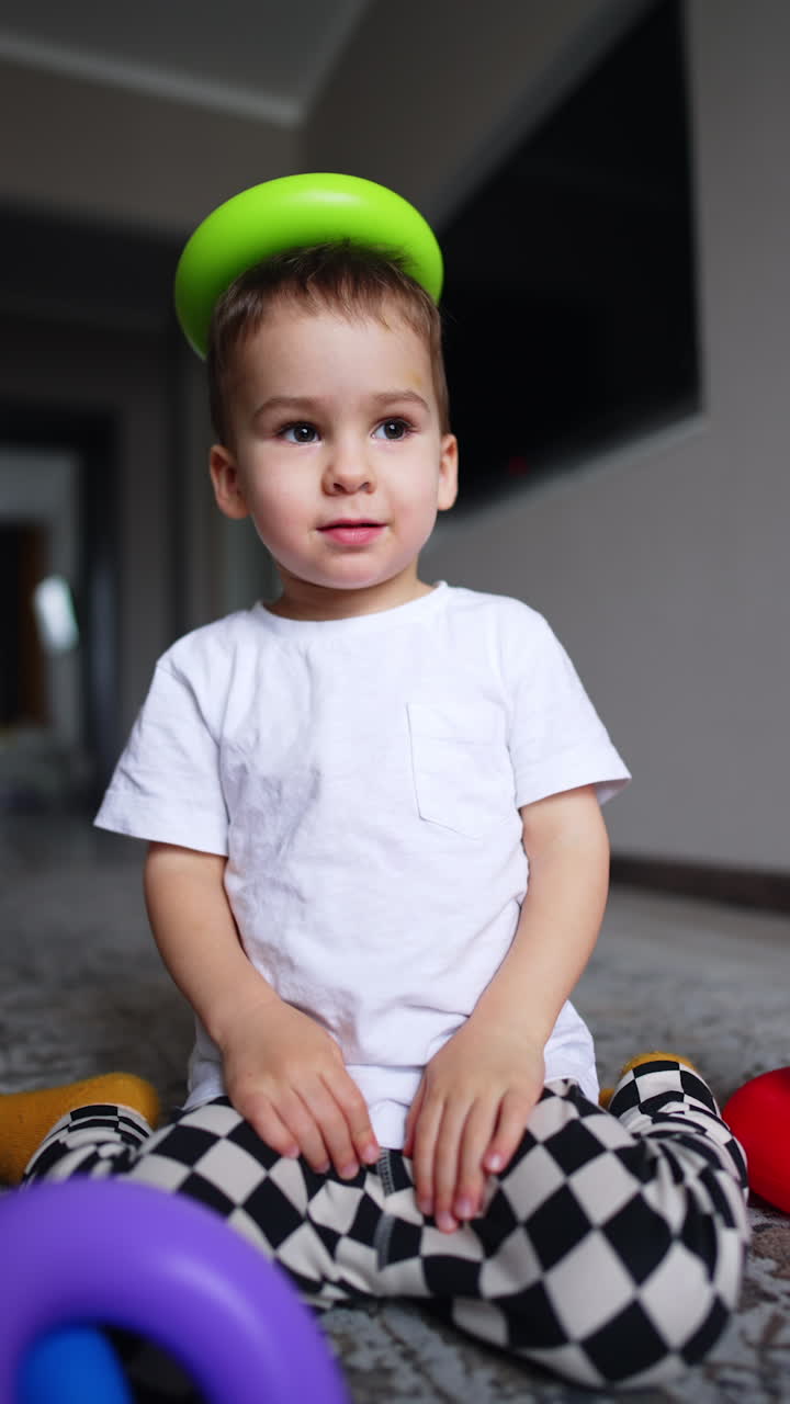 Adorable Caucasian baby sits on the floor with a pyramid detail on his head. Low angle view on the child playing at home. Vertical video.