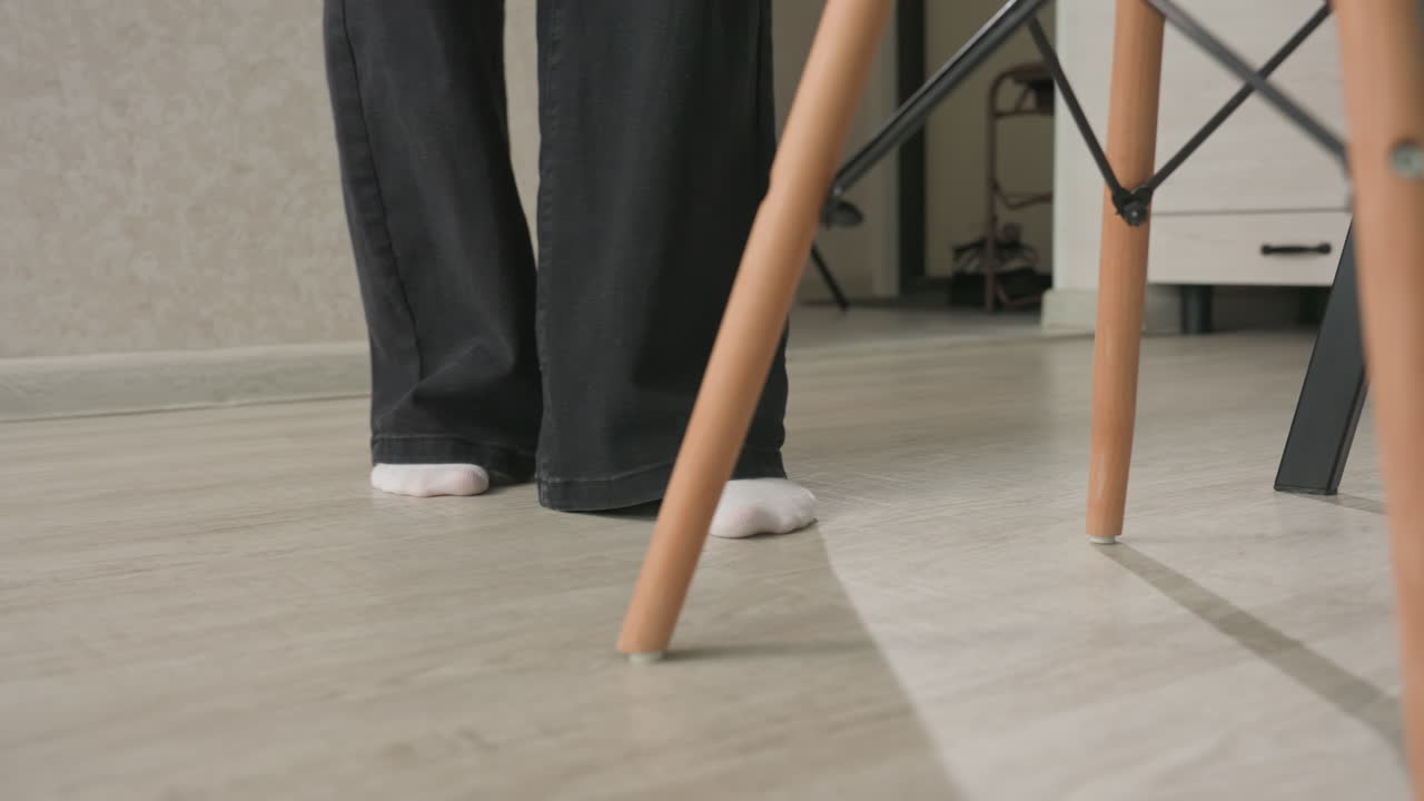 close up of two adults legs walking away from doorway into living room near sofa, one holding white handbag with pink trim, barefoot and sock contrasting on wood floor
