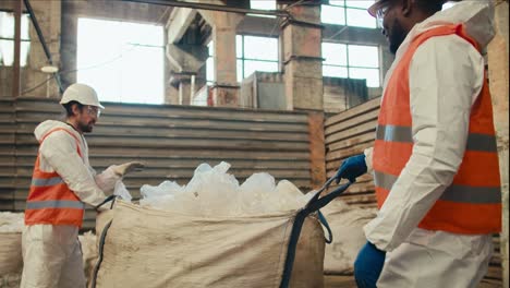 A-man-with-Black-skin-in-a-white-special-uniform-and-an-orange-vest-lays-out-and-sorts-cellophane-garbage-together-with-his-colleague-a-man-with-a-beard-in-a-white-helmet-at-a-waste-processing-and-sorting-plant