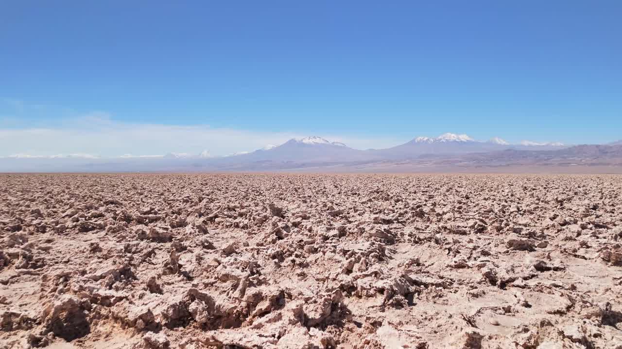 Low-altitude drone footage over the Atacama Desert salt flats during daylight. The drone moves forward, capturing salt crusts, saline soil, lithium, and snow-capped mountains on the horizon.