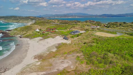 A beautiful aerial shot slowly pushes in over a stunning Japanese coastline, featuring a sandy beach, turquoise sea with gentle waves, and vibrant green hills under a partly cloudy sky