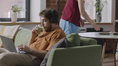 chico caucásico trabajando en una computadora portátil sentado en el sofá en un apartamento compartido mientras su compañera de cuarto le da un vaso de café