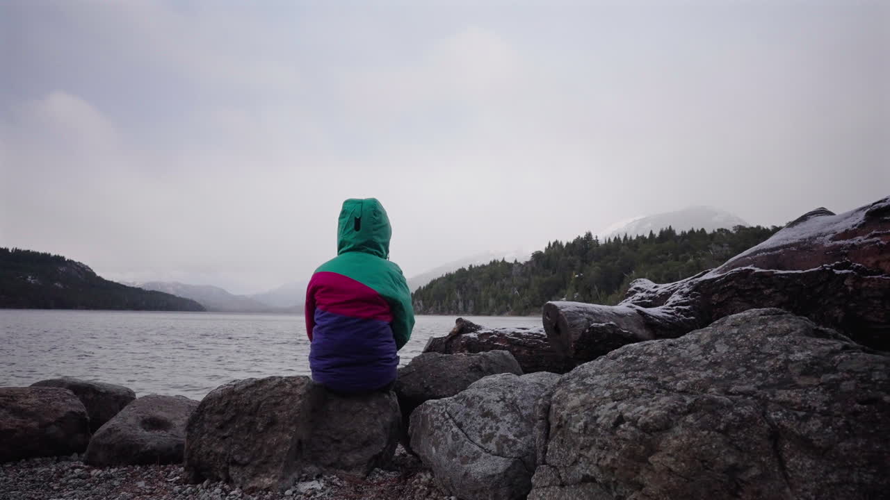 Young girl in colorful jacket sits alone by cold, grey Patagonian lake, deep emotional stillness and introspective calm
