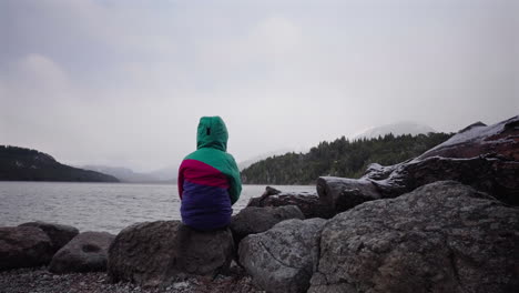 young girl in colorful jacket sits alone by cold, grey patagonian lake, deep emotional stillness and introspective calm