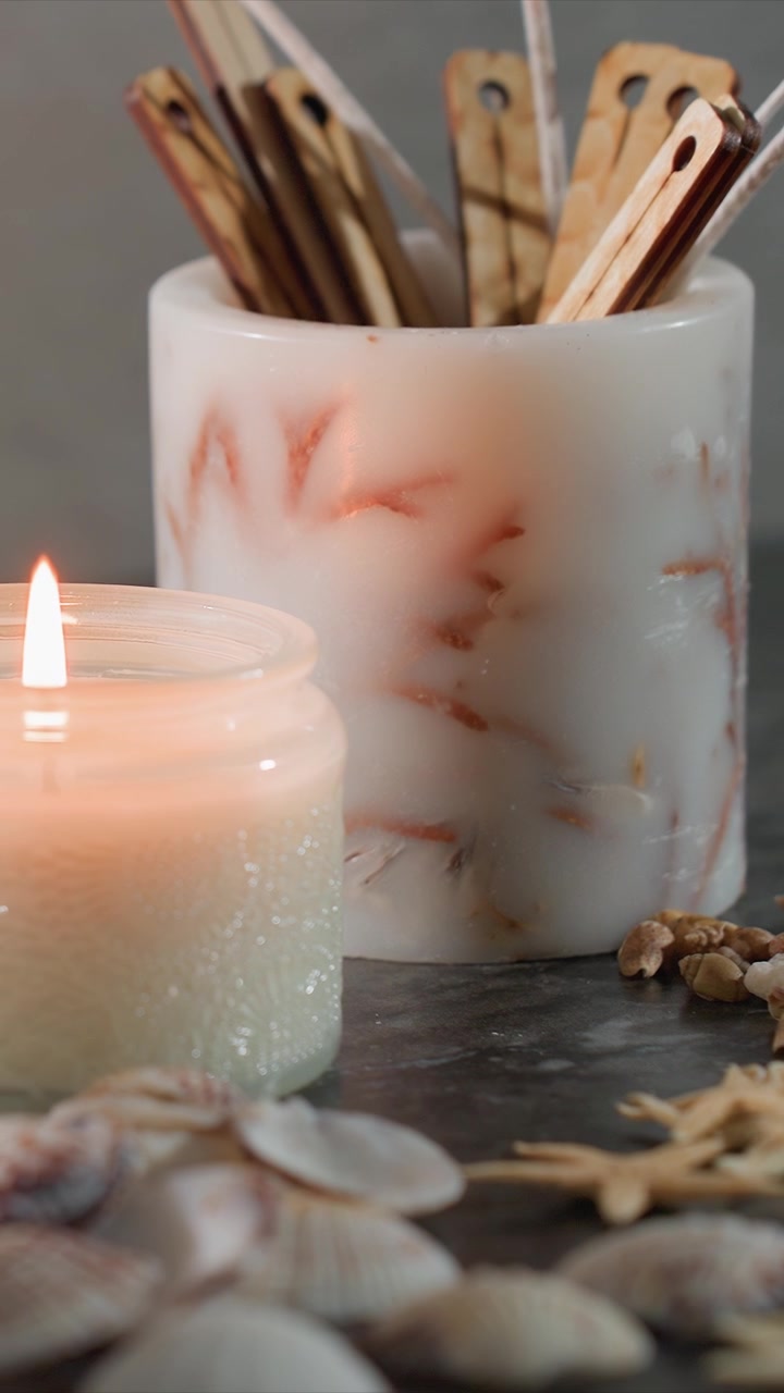 Primo piano di una candela accesa e di una candela a colonna decorativa con oggetti a tema marino come conchiglie, stelle marine e bastoncini di legno disposti su una superficie scura. L'illuminazione soffusa crea un'atmosfera calda con sottili