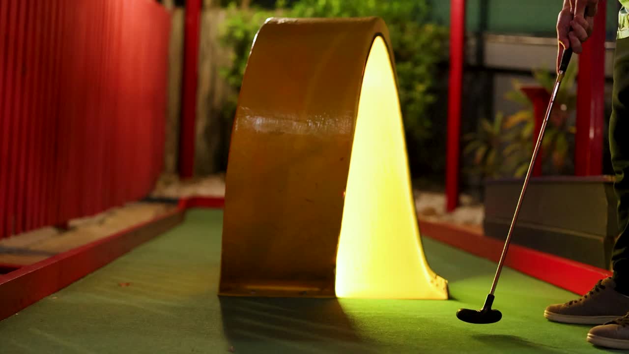 A player attempts a mini golf putt through a glowing tunnel in a colorful indoor course with ambient lighting
