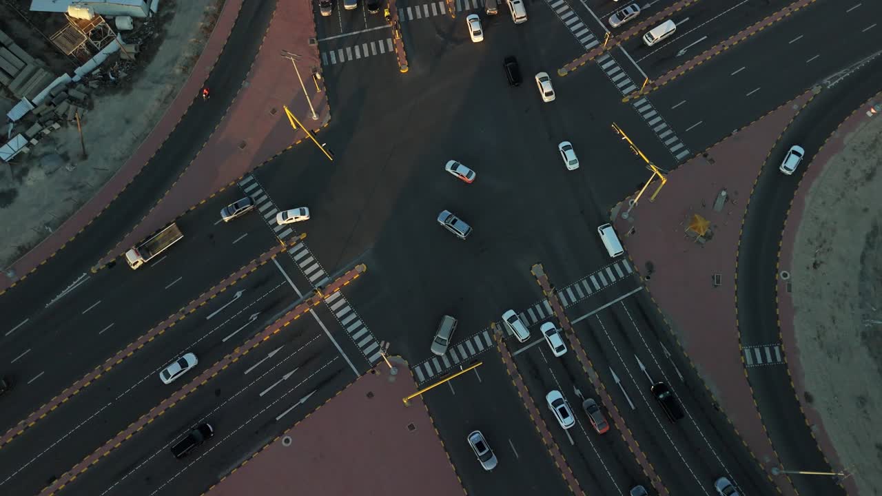 Top-down drone over a busy multilane intersection at golden hour—cars, zebra crossings, and crisp lane markings forming a geometric city-grid pattern with long shadows in a Gulf-region urban setting