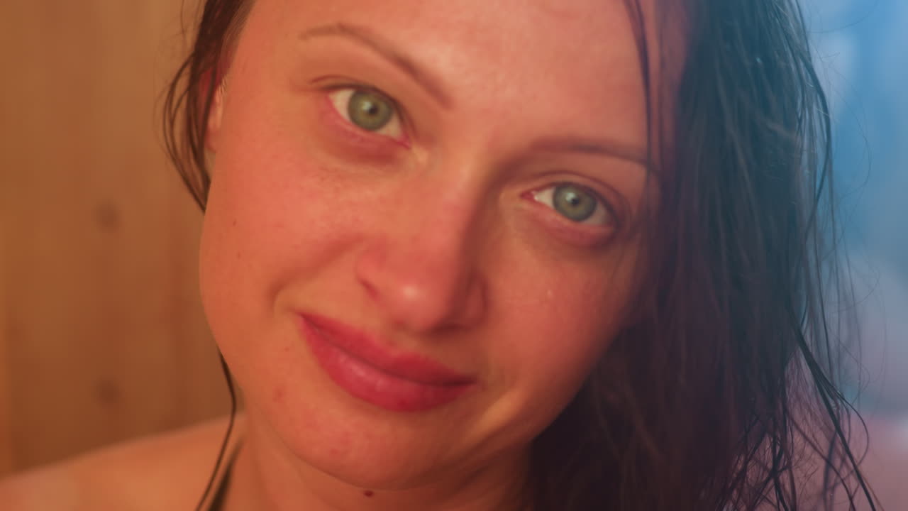 two women wrapped in towels relax inside smoky sauna room, one gazing upward with serene green eyes and soft smile through mist, damp hair framing calm expression against warm wooden walls