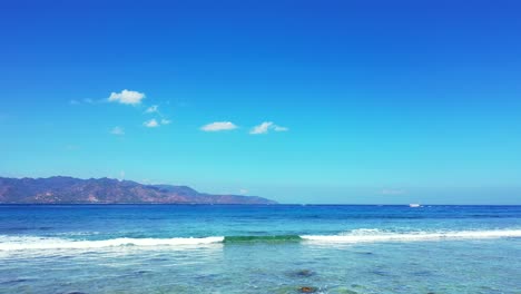 Paradise-seascape-with-white-waves-foaming-over-rocky-seabed-on-seashore-of-tropical-island-with-light-blue-sky-background-in-Bali,-copy-space