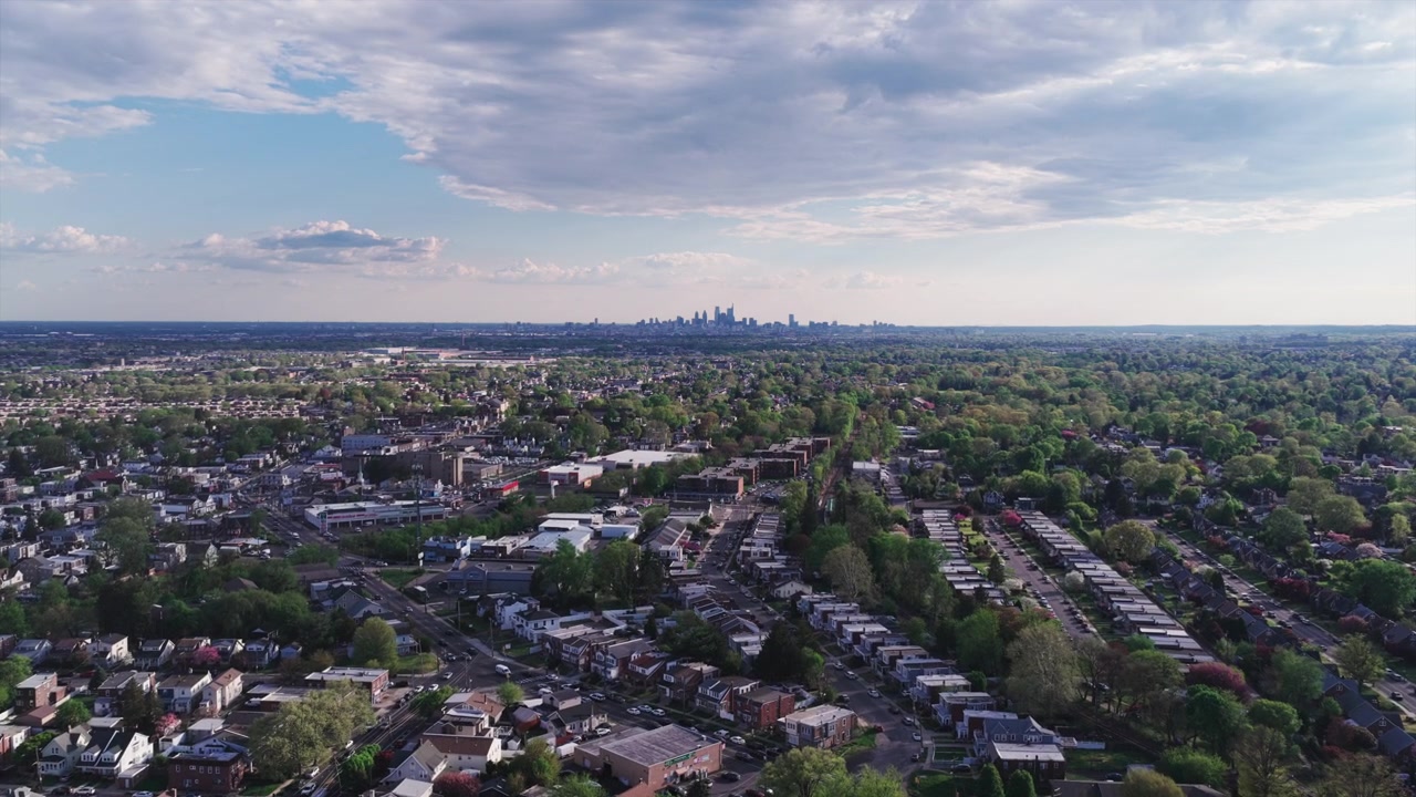 Wide aerial drone view of a lush Philadelphia suburban landscape stretching toward the faint downtown skyline on the horizon under a partly cloudy spring sky.