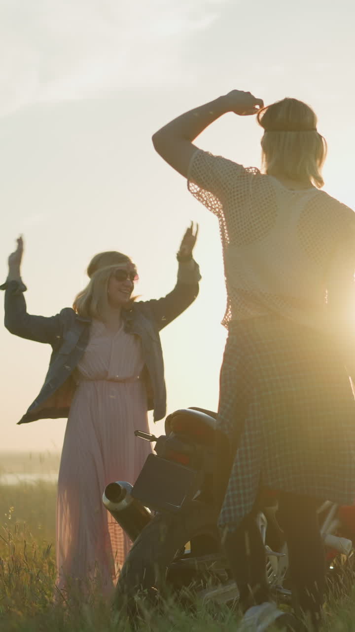 Two women, wearing headbands and sunglasses, dance joyfully in an open grassy field near a motorcycle at sunset