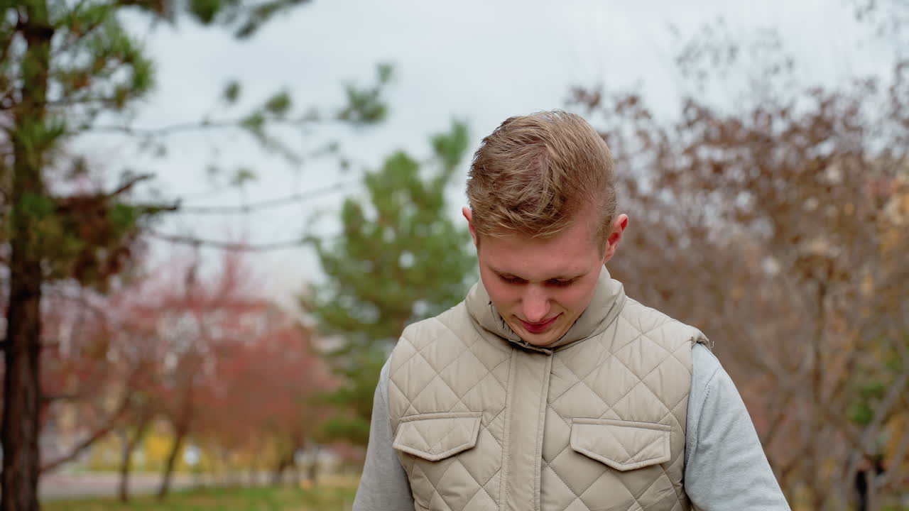 Focus boy in jacket walking outdoors as wind blows, bending head slightly then lifting it up with trees swaying in background, surrounded by autumn colors in peaceful urban setting on cool breezy day