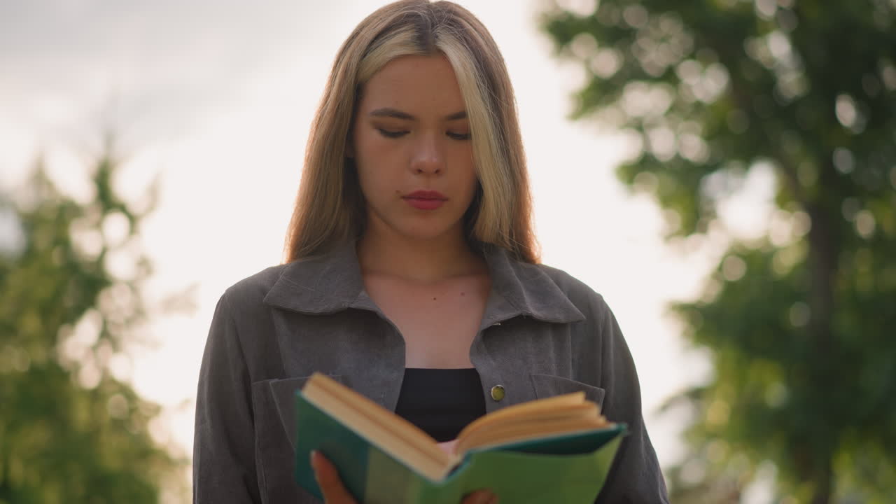 dama con ropa gris de pie al aire libre, cabeza ligeramente inclinada, hojeando las páginas de un libro con la luz del sol reflejándose suavemente en el fondo características borrosos árboles y edificios