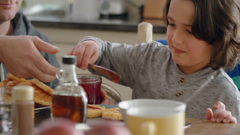 Fröhlicher-Kleiner-Junge,-Der-Zum-Frühstück-Frische-Waffeln-Isst-Und-Köstliche-Hausgemachte-Leckereien-Genießt,-Während-Sein-Vater-Seinem-Sohn-Zu-Hause-In-Der-Küche-Hilft-4k