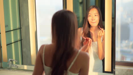 Hübsche-Asiatische-Frau-Vor-Dem-Badezimmerspiegel,-Die-An-Einem-Sonnigen-Morgen-Lippenstift-Auf-Die-Lippen-Aufträgt