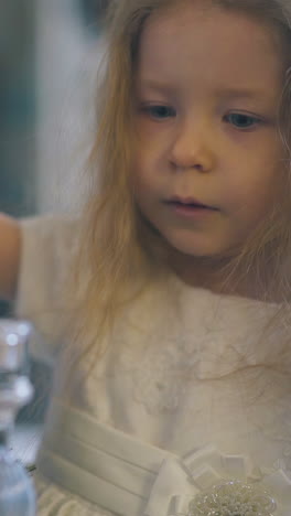 adorable little blonde girl in festive dress looks at decorative bird cage with fir branches and toy in light room