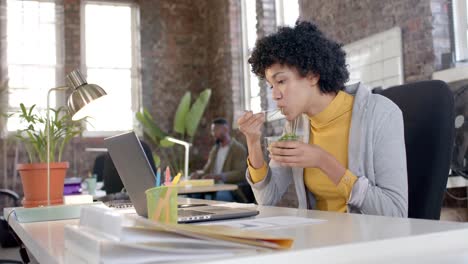 Focused-biracial-casual-businesswoman-using-laptop-and-having-lunch-in-office-in-slow-motion