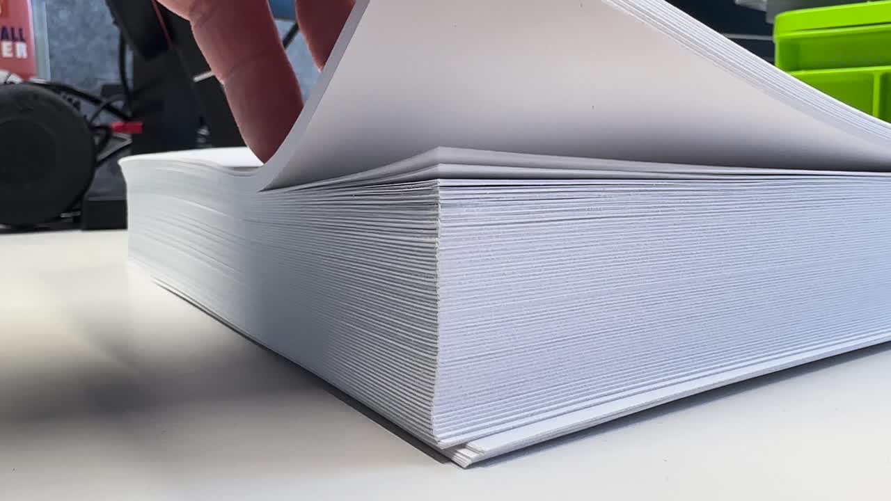 Close-up of a hand flipping through a thick stack of white paper on a desk, highlighting office work, documentation, printing, and everyday business routine in a clean workspace