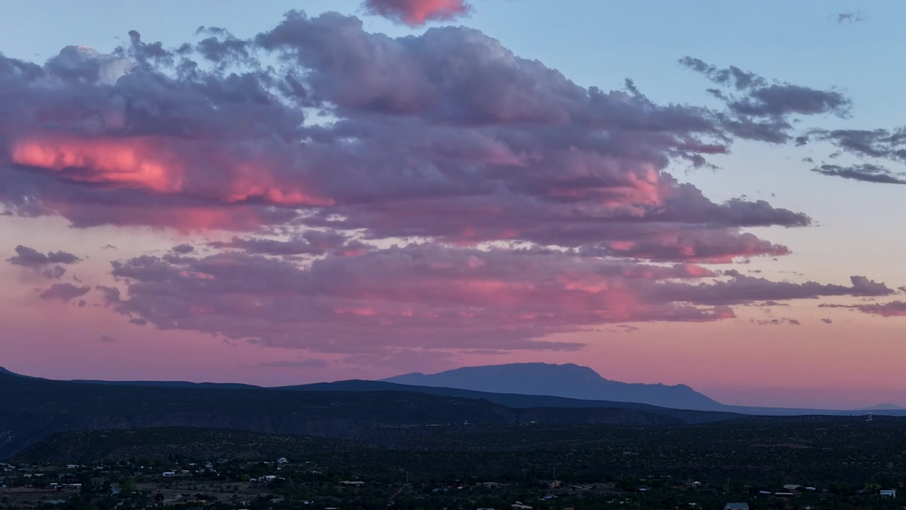 Timelapse con drone 4K UHD che cattura un tramonto vibrante. Nuvole morbide e strutturate cambiano dal rosa brillante al viola intenso sopra una vasta città desertica e le lontane Montagne Sandia nel Nuovo Messico.