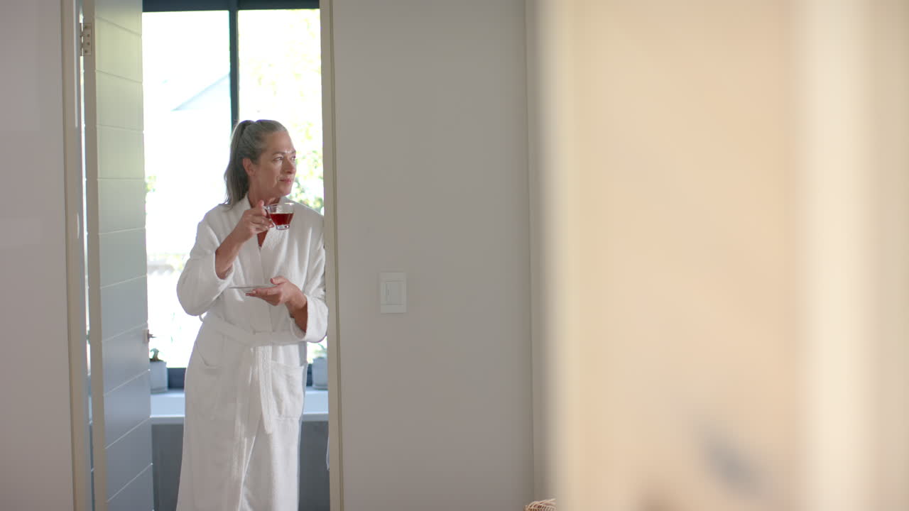 Holding cup of tea, mature woman in bathrobe standing in doorway at home