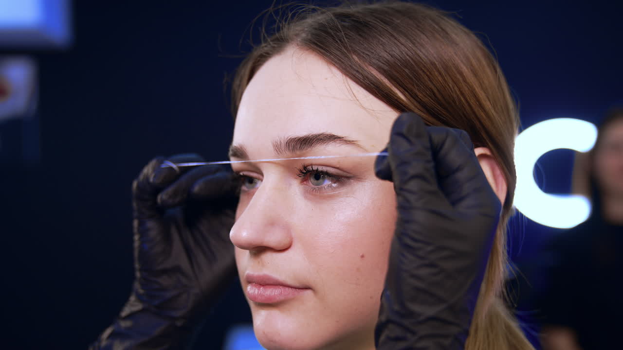 Young pretty woman face. Hands in black latex gloves attach the thread to the girl's forehead in order to mark the lines for eyebrow make up. Close up.