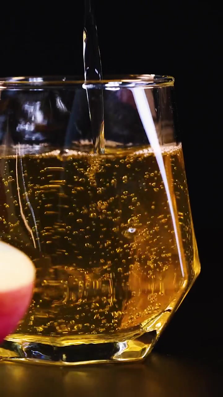 Jus apel keemasan mengalir ke dalam gelas geometris dengan latar belakang gelap dan gelembung