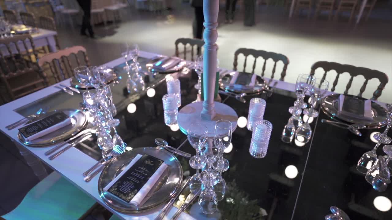 A detailed shot of an elegant banquet table set for a wedding or gala. The setup features silver charger plates, crystal glassware, and a black, reflective tabletop
