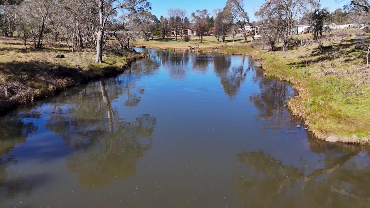 Drone camera glides above a calm stream with ducks, passing through grassy banks and residential houses under bright daylight and clear blue sky