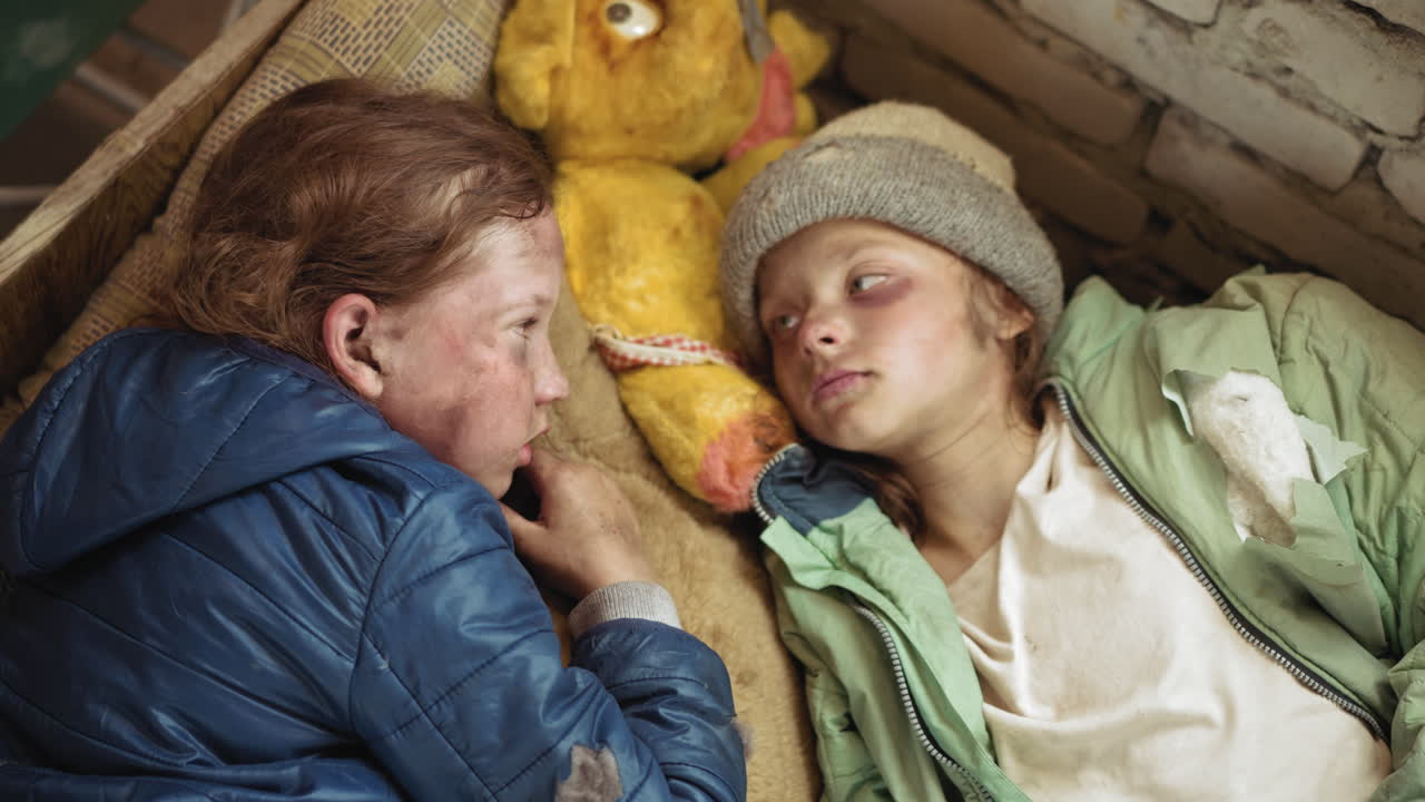 Adolescents lying close on mattress with worn stuffed toy between them, gazing silently with weary eyes, faces marked by dirt and exhaustion, torn jackets and rough wall