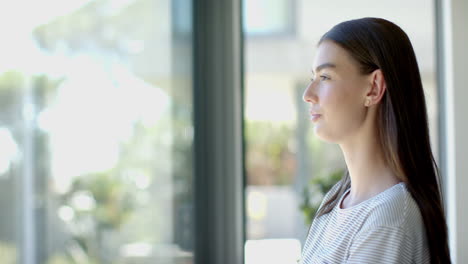 Caucasian-young-woman-standing-by-window,-looking-thoughtful