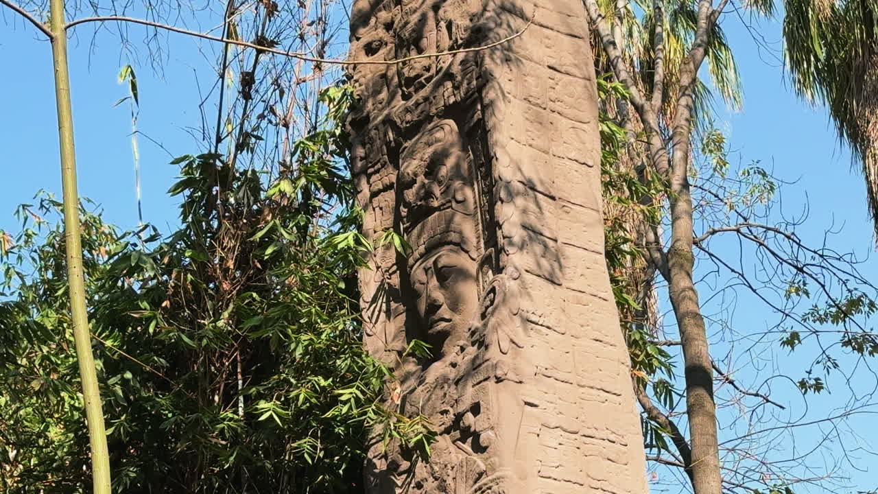 Imponente colonna di pietra scolpita con un antico rilievo facciale al Museo Nacional de Antropología a Città del Messico. Scultura precolombiana circondata da alberi tropicali sotto un cielo azzurro.