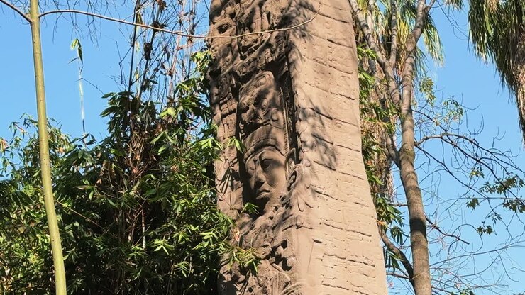 Imponente colonna di pietra scolpita con un antico rilievo facciale al Museo Nacional de Antropología a Città del Messico. Scultura precolombiana circondata da alberi tropicali sotto un cielo azzurro.