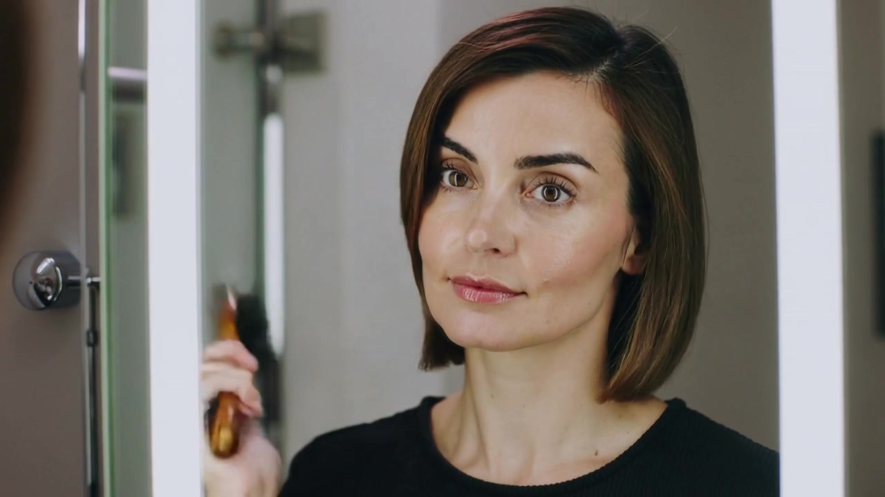 Una donna che si spazzola i capelli controlla il suo riflesso in uno specchio LED mentre sistema il caschetto con una spazzola piatta in legno, preparandosi per la giornata.