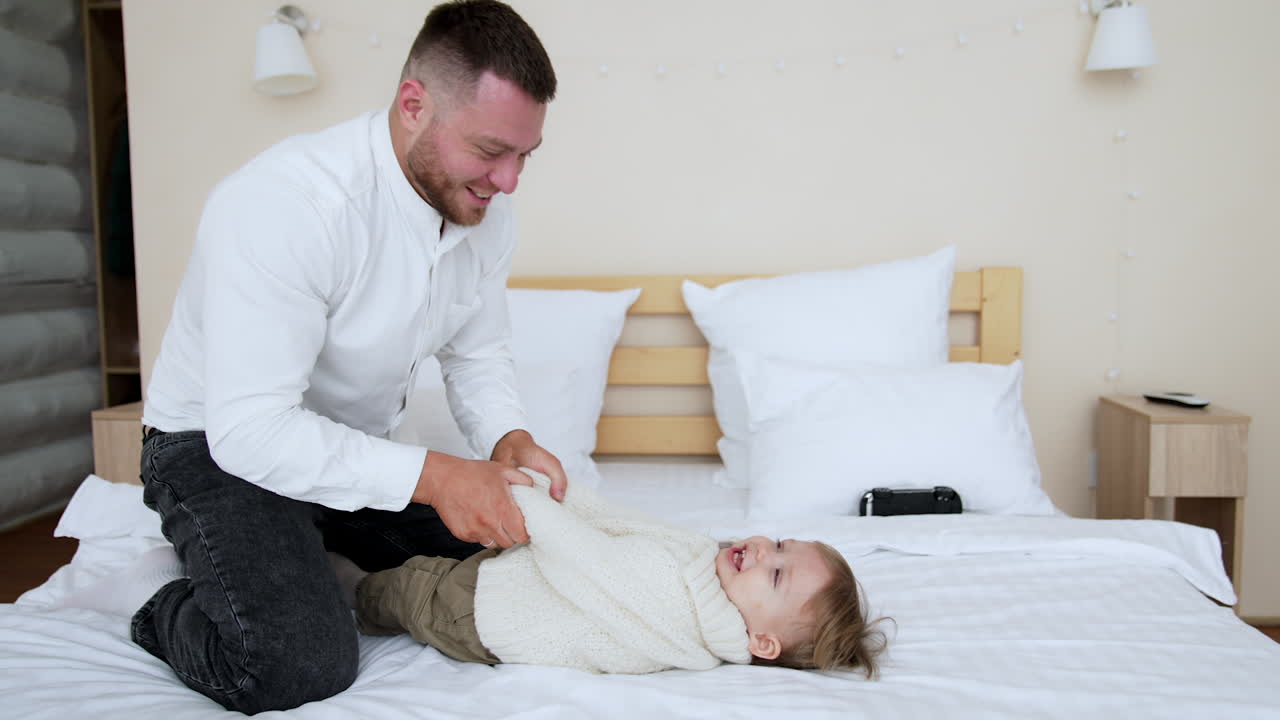 Dad and his toddler son are playing on the big bed. Loving father kisses his baby boy on the cheek and raises him above the head.