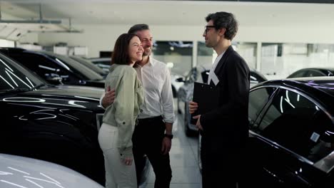 Happy-couple-a-middle-aged-man-with-gray-hair-together-with-his-brunette-wife-is-having-fun-talking-with-a-car-dealership-assistant-in-a-business-suit-about-cars-in-a-car-showroom