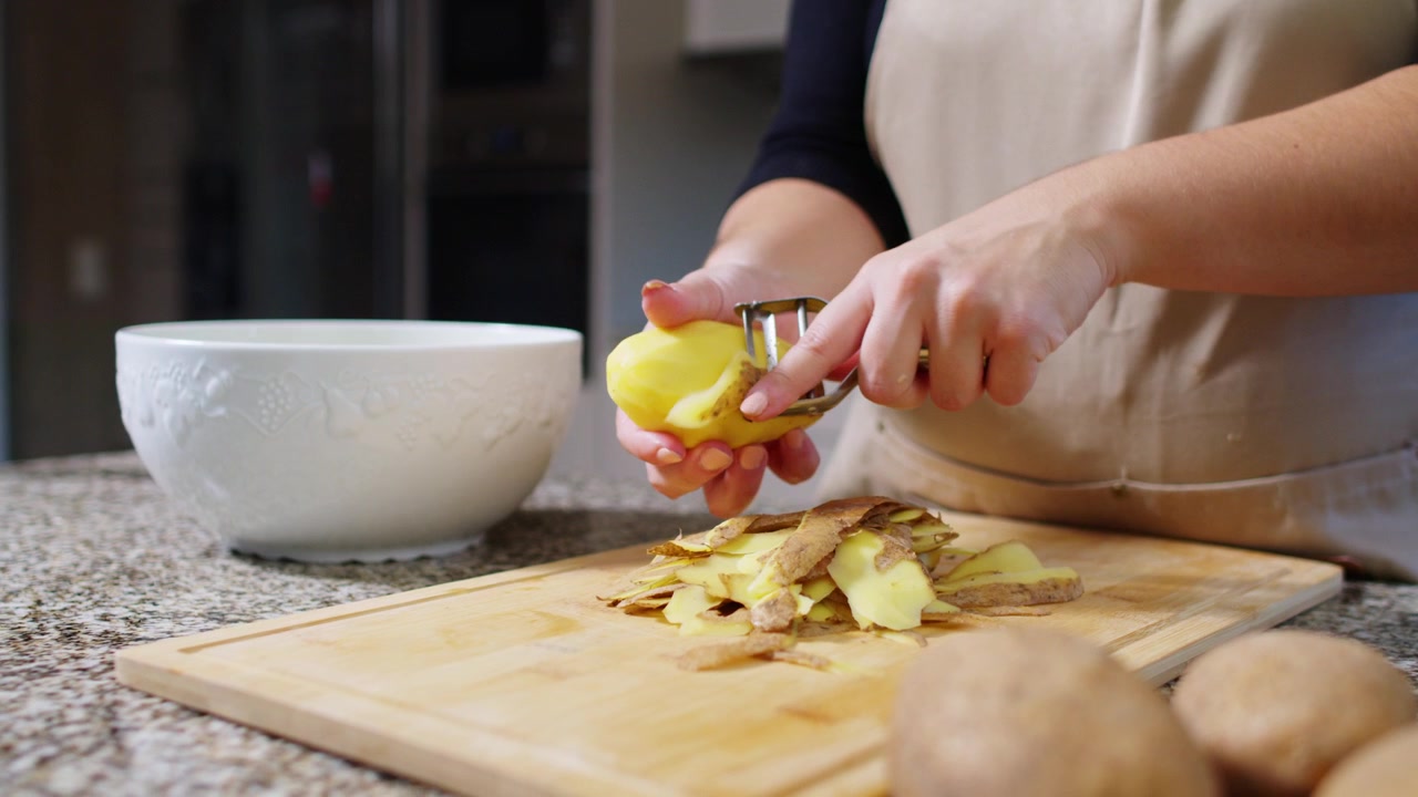 Ripresa al rallentatore di lato di una donna che pela patate con un pelapatate, con una vista più ampia della cucina sullo sfondo.