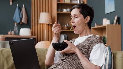 mature woman eating from a bowl with chopsticks while looking at a laptop on a sofa at home