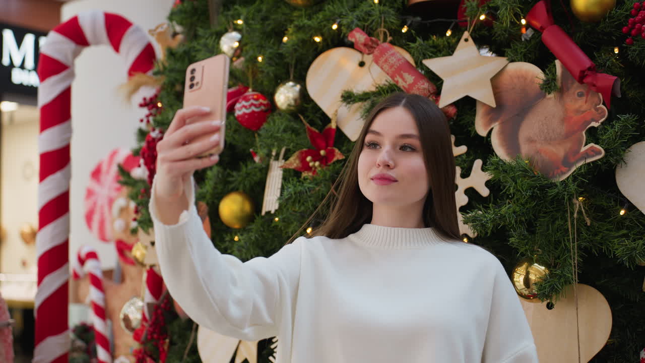 Beautiful woman in white sweater taking a festive selfie in front of large decorated Christmas tree with twinkling lights, wooden ornaments, and holiday decorations in a cheerful setting