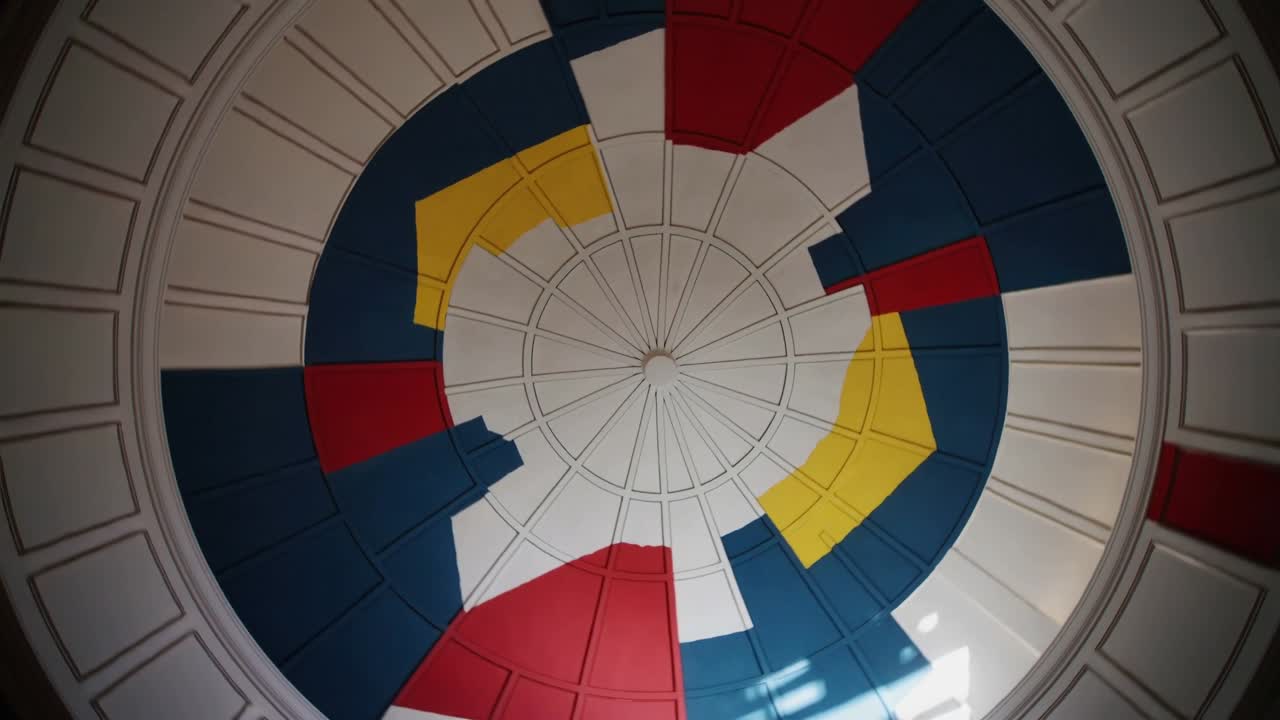 Telecamera rotante che mostra un rosone a cupola nel soffitto di una rotonda, rivelando pannelli colorati e una chiazza di sole