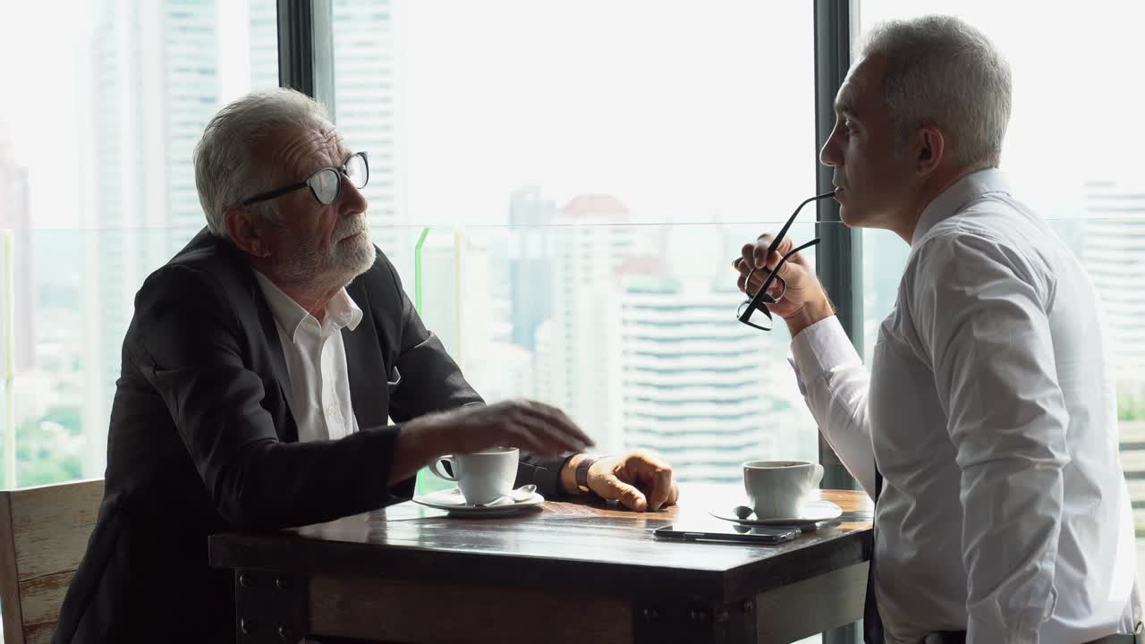 two senior businessman take a break drinking coffee and discussing business strategy during break on table in coffee shop at modern restaurant on a tall building cityscape background
