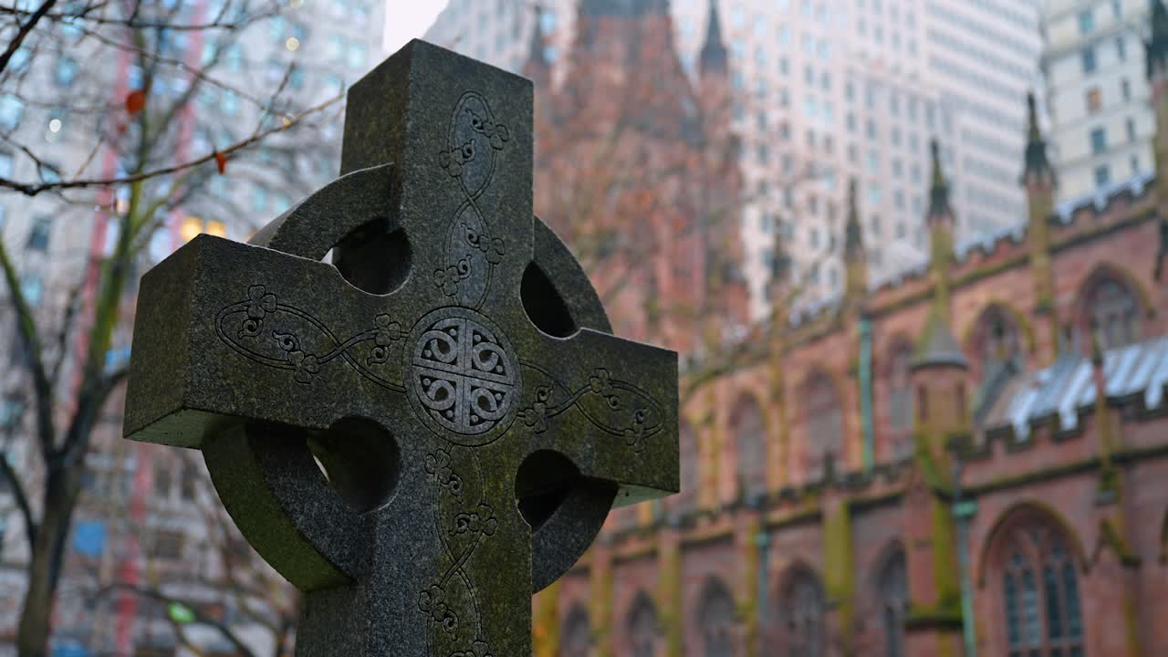 Nærbillede af en keltisk gravstenskors i sten på en kirkegård. Detaljeret billede af et indgraveret stenkors med en historisk kirke og bybygninger i en sløret baggrund.