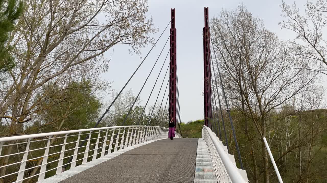filming on a modern bridge with two colors of rust red on the two pillars and white on the walkways where we see how a woman with a violet skirt and a black shirt crosses it, enjoying the landscape