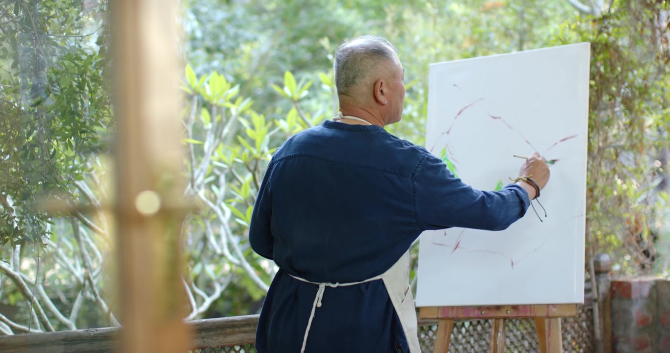 Uomo anziano asiatico di fronte a un cavalletto sulla veranda, che dipinge di verde con un pennello, dando forma al disegno, indossando un grembiule