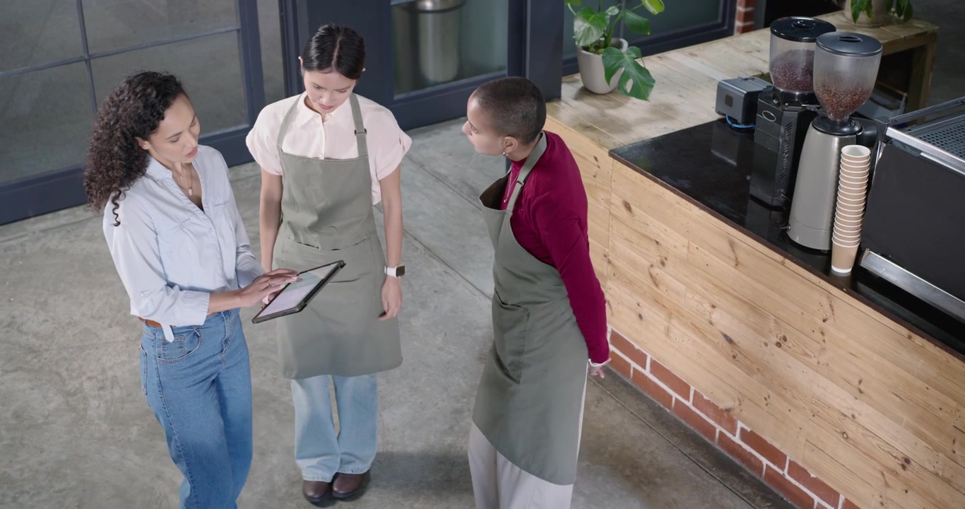 Gerente sosteniendo una tablet acercándose al mostrador mostrando la pantalla para una reunión informativa mientras el personal femenino asiente