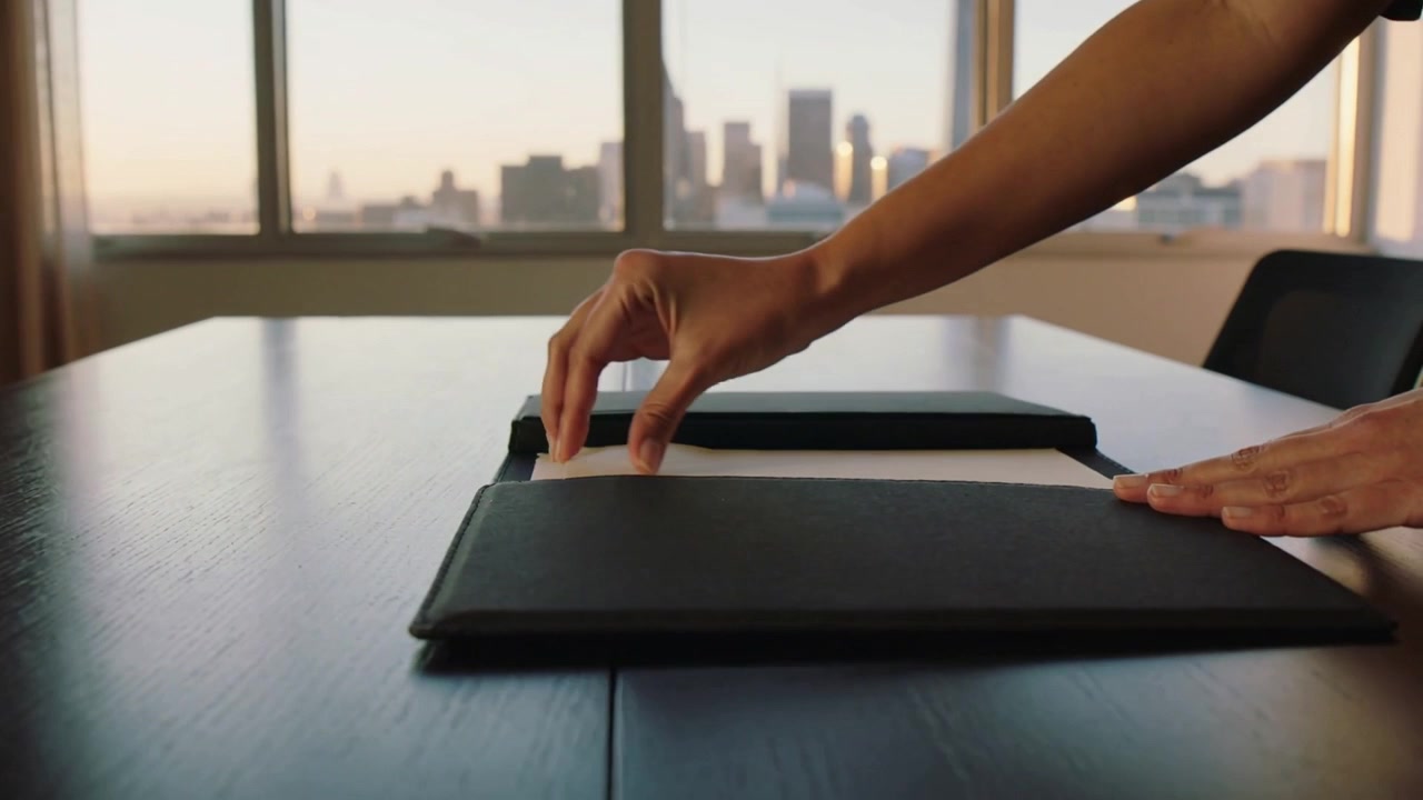 Placing bare arms smoothing tucking pale envelope on black leather folio filing by window skyline